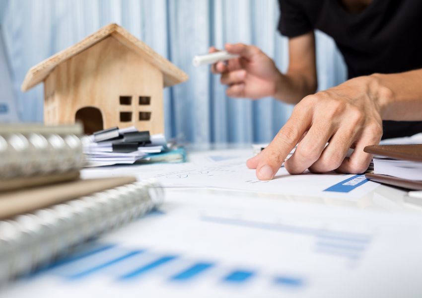 hand pointing to stats and figures on a paper with a home figurine in the background