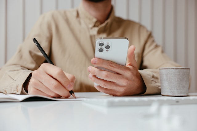 person in a beige shirt holding a cell phone and taking notes on a piece of paper