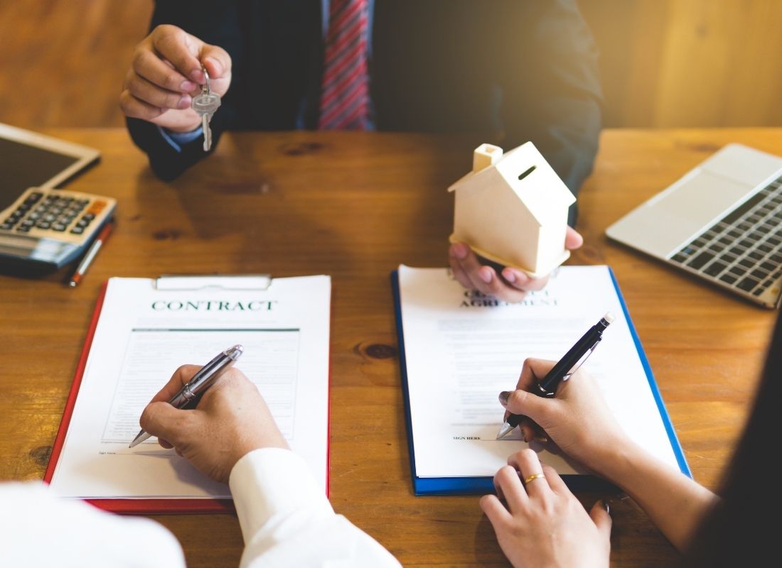 someone holding house figurine in one hand and keys in the other as two people sign contacts