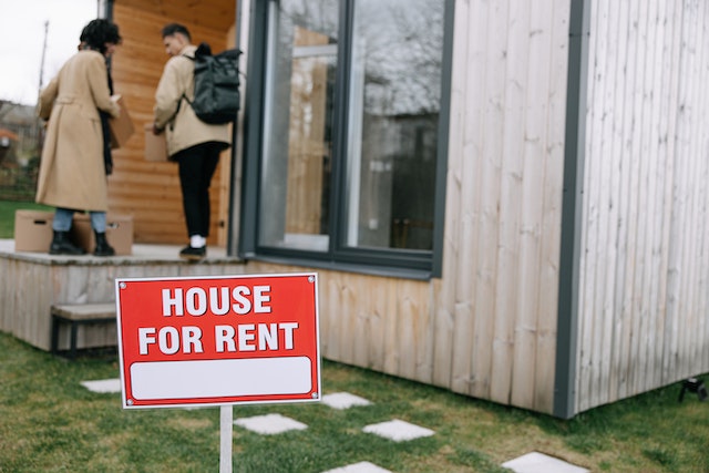 two people moving boxes into a house with a sign that says house for rent in front